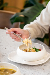 A close-up of a hand holding a fork with pasta, served in a modern white bowl. Fresh vegetables and herbs are visible, emphasizing a healthy and appetizing meal.