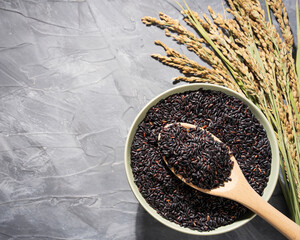 A bowl of black-rice on a gray table next to a wooden spoonful and ears of rice.Bowl is filled with...