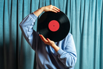 Woman in  blue sweatshirt covers her face with vinyl record on blue velvet curtains background