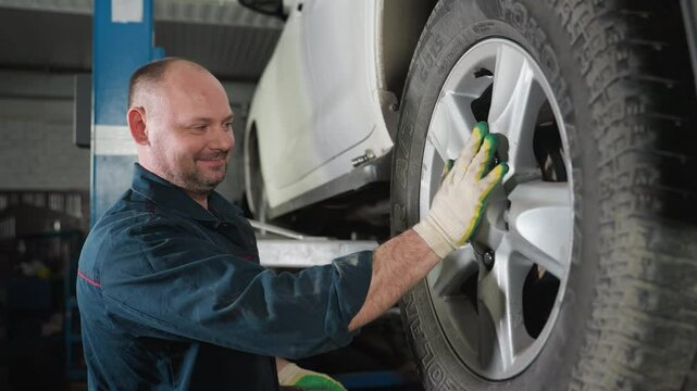 Mechanic in blue uniform with hairy hand and white gloves smiling while adjusting and aligning car tire in auto repair workshop, ensuring proper positioning and safety