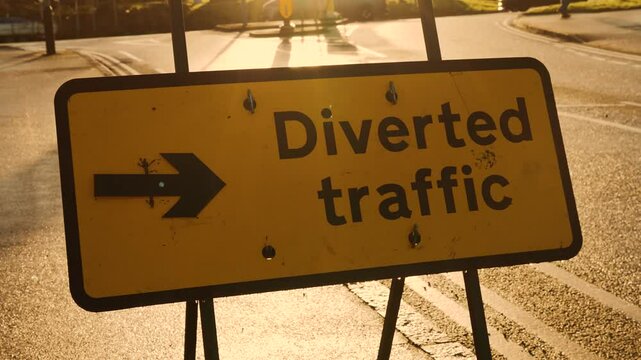 Detour sign standing on sunlit road. Highway sign contour in morning glow. Traffic redirection board appearing as dark figure in bright sunrise rays