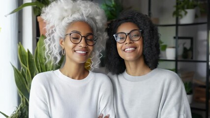 Two women share laughter and warmth in a bright indoor green space, showcasing their unique styles and the bond of friendship while surrounded by nature's beauty.