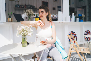 Young woman in activewear sitting at an outdoor cafe, checking her smartwatch while holding a smartphone. She has a yoga mat and water bottle nearby, enjoying a calm break after exercise