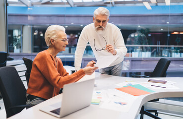 Obraz premium Senior business professionals reviewing documents together at a modern office table, man leaning forward attentively while woman holds paperwork. Concept of teamwork, experience,and strategic planning