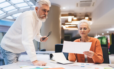 Fototapeta premium Senior business professionals reviewing documents together at modern office, man watching while holding glasses. Concept of collaboration, experience, focused decision-making in corporate planning
