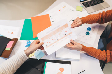 Close-up, colleagues reviewing printed chart at meeting. Various documents, colorful folders, and sticky notes scattered on table, organized yet active workspace focused on data analysis and planning