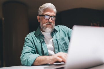 Senior man with white beard and glasses, intently focused on his laptop in a modern office. Wearing...