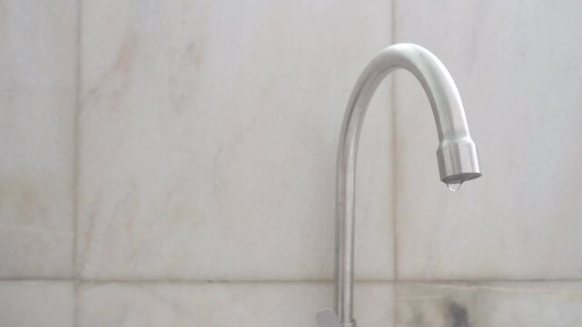 A stainless steel silver faucet tap dripping water with white marble wall at background