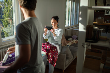 Woman taking picture of boyfriend ironing clothes at home