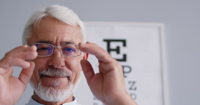 Happy senior man puts on new eyeglasses Closeup face handsome white haired bearded old man in glasses looking at camera with blurred optometrist's ophthalmologist's visual eye exam chart in background