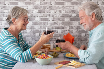 Cheerful caucasian senior couple toasting with red wineglass while sitting face to face at table having brunch or breakfast together at home