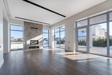 Spacious living room with panoramic windows in a luxurious mansion. Parquet floor, fireplace decorated with beige marble tiles. Unfurnished.