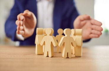 Businessman in suit holding hands above wooden toy people figures standing in circle at desk protecting human rights. Society, people community care and safety concept in business.