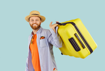 Young funny happy smiling man tourist in beach hat standing with suitcase, looking at camera going on summer holiday trip isolated on blue studio background. Travel and vacation concept.