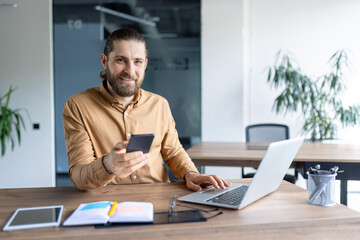 A bearded man sits at a desk in a contemporary office space, utilizing a smartphone and a laptop. Various tools and notes are present symbolizing efficiency, work, and communication.