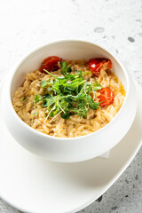 A creamy orzo dish garnished with fresh microgreens and cherry tomatoes in a white bowl, placed on a light textured surface.