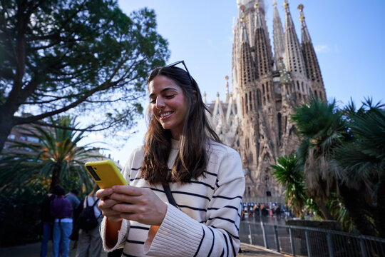 Cheerful young woman using mobile phone in front of the sagrada familia basilica, enjoying her travel experience connected to internet. Copy space.