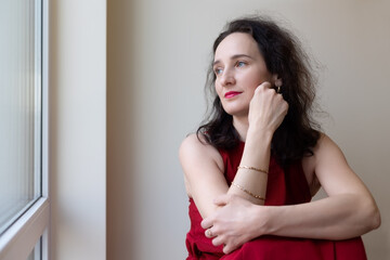 Thoughtful woman in red dress sitting by the window, gazing outside with a serene expression.