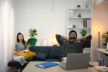 Young businessman working at home office on laptop computer, finishing his online work, while his girlfriend and roommate sitting behind on sofa relaxing and waiting. Freelance business. couple.