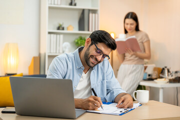 Young businessman working at home office on laptop computer, finishing his online work, while his girlfriend and roommate sitting behind on sofa relaxing and waiting. Freelance business. couple.