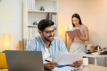 Young businessman working at home office on laptop computer, finishing his online work, while his...
