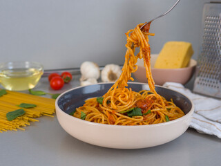 Balanesi pasta with tomato sauce and basil is being lifted by a fork from a bowl, with fresh ingredients like tomatoes, garlic, olive oil and cheese visible on a kitchen counter