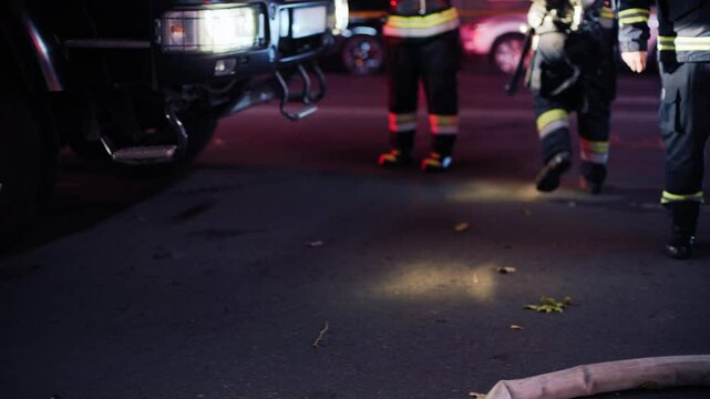 A team of three firefighters with full protective gear, walks through an emergency scene near firetrucks, preparing to respond.