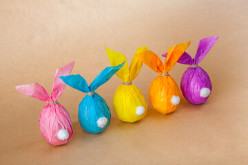 Five eggs wrapped in colored paper resemble adorable bunnies with ears and white tails. cute bunnies stand on a light brown surface, creating a charming Easter scene.