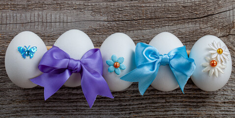 Several eggs are on the wooden table prepared for the Easter holiday. Each egg is decorated with colorful beads, as well as butterflies and stars made of fabric. Some eggs are adorned with blue ribbon