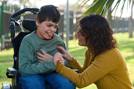 Mother tickling son with disability sitting in wheelchair in park - cerebral palsy concept