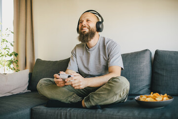 Happy man playing video game and eating snacks, sitting in a cozy living room. © polinaloves
