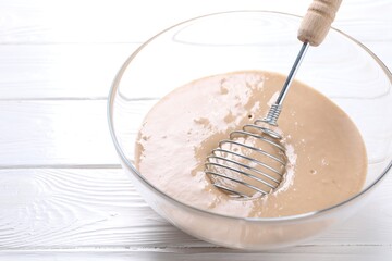 Whisk and bowl of dough on white wooden table, closeup. Space for text