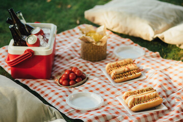 Picnic with hot dogs and cold drinks in cooler bag on red tablecloth in a park.