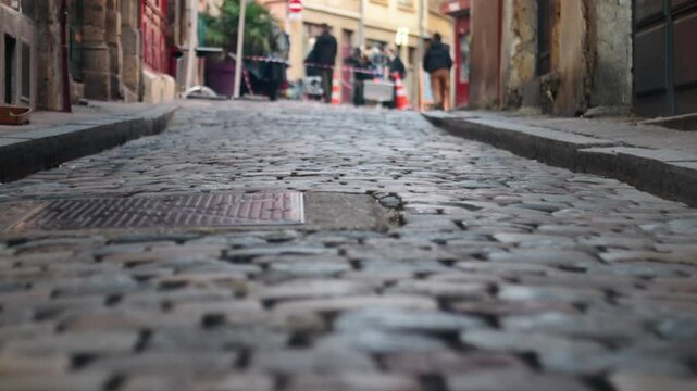 Focus changes on a cobblestone street in lyon, france, with a metal grate and blurred people in background