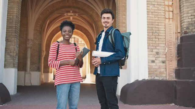 Students standing in front of big building. Girl in red shirt holding notes and male holding architectural drawing rolled into paper tube. Glancing at camera while smiling. Wearing backpacks.