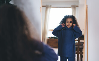 African American girl with curly hair standing in front of mirror, adjusting her blue turtleneck outfit. She looks at her reflection with confident and thoughtful expression in bright, cozy bedroom.