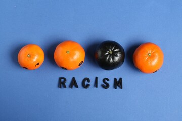 One black tangerine among orange ones and word Racism made with beads on blue background, top view