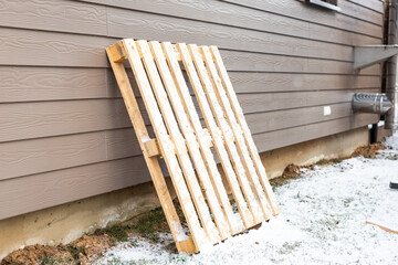 A wooden pallet rests on the ground near a building with horizontal siding. Snow covers parts of the ground, revealing patches of grass and soil.