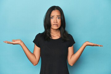 Filipina young woman on blue studio confused and doubtful shrugging shoulders to hold a copy space.