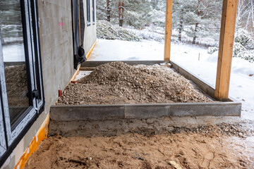 Outdoor construction site with a rectangular foundation, gravel pile, wooden posts, and snow covered ground surrounded by trees in a wintry setting.