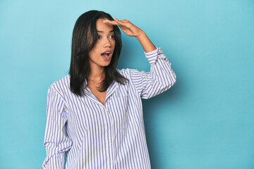 Filipina young woman on blue studio looking far away keeping hand on forehead.