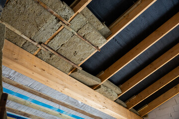 Exposed wooden beams and insulation material are visible in a building under construction, with a black vapor barrier on the ceiling section.