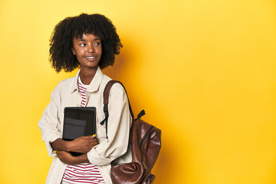 Tech-savvy African-American teen girl with tablet and backpack on yellow studio backdrop.