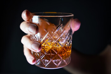 A close-up of a man's hand holding a half-full crystal glass with whiskey or rum in bright gold color, illuminated on a dark background.