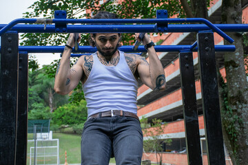 Young tattooed man doing pull-ups and parallel bars exercise in a public park surrounded by nature