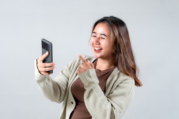 Fototapeta premium A young woman smiles and points at her smartphone, expressing joy and excitement while engaging with her device in a neutral background.