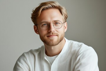 Fototapeta premium Young man with glasses smiling in casual white attire against a neutral background