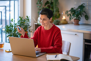 Relaxed mature woman smiling and making online purchases, browsing marketplaces, booking travel, ordering delivery with credit card. Preparing for vacation, enjoying electronic commerce convenience