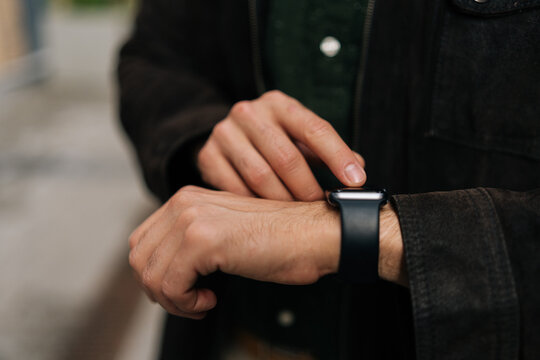 Close-up hands of unrecognizable man interacting with smartwatch, checking time, reading notifications and navigating apps while standing outdoors in urban setting. Concept of modern city lifestyle.