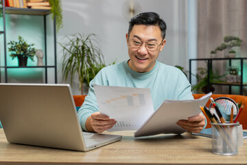 Happy Asian man freelancer work with documents at home analyzing information read business papers...